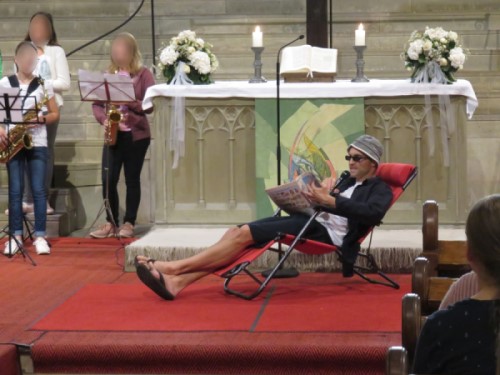 Schulanfangsgottesdienst. Mann mit Hut, kurzer Hose, Sonnenbrille und Zeitung liegt in einem Sonnenstuhl vor dem Altar der Stiftskirche