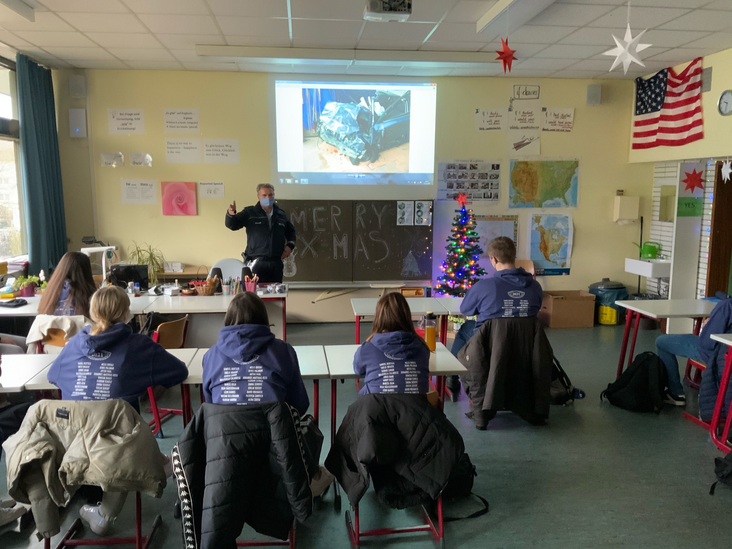 Verkehrserziehung durch einen Polizisten im Klassenzimmer mit einer sitzenden Schülergruppe. Über der Tafel wird ein zerstörtes Auto projiziert.