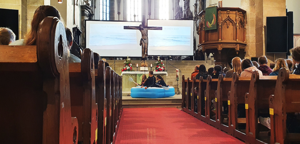 SchÃ¼ler in der Kirche, im Hintergrund zwei LeinwÃ¤nde, ein Kreuz und eine SchÃ¼lergruppe in einem Schlauchboot
